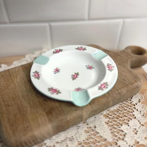 May include: White ceramic ashtray with a floral rose pattern. The ashtray has a round shape with three notches and a light blue accent. The roses are pink and green. The ashtray is on a wooden cutting board.