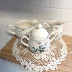 May include: A cream-colored ceramic tea set with floral designs, including a sugar bowl with a lid, and two cream pitchers. The set features pink and blue floral accents and a delicate blue trim. The set is placed on a wooden board with a lace doily.