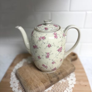 May include: Cream-colored ceramic teapot with a lid, adorned with a delicate pattern of pink roses and green vines. The teapot has a curved spout and handle, and sits on a wooden cutting board. The rim and handle have gold trim.