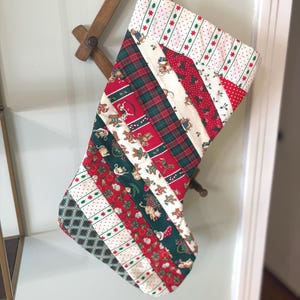 May include: A Christmas stocking constructed from diagonal strips of fabric featuring various holiday patterns including gingerbread figures, red and green plaid, white with red and green dots, and dark green geometric prints with a white hanging loop.