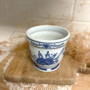 May include: A white ceramic planter featuring a blue floral pattern painted around the exterior. The vessel has a rounded body, a flared rim with a thin blue line, and a circular base. The design includes detailed blue leaves and blossoms.