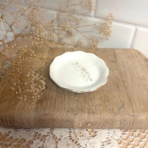 May include: A small, round, off-white ceramic dish with a scalloped edge and a textured surface. The center of the dish features a faint, light brown floral imprint. The rim of the dish is finished with a thin, metallic gold-colored trim.