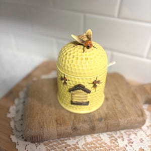 May include: A yellow ceramic honey pot shaped like a beehive, with a textured surface and a small bee figurine on top. The pot has a small wooden-looking house design on the front and several bee illustrations. A small spoon rests on the side.