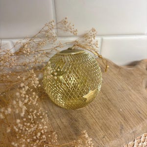 May include: A spherical gold-colored metal ornament featuring a fine wire mesh grid pattern and small attached star-shaped accents. The ornament includes a metallic cap at the top with a thin gold string loop for hanging purposes.