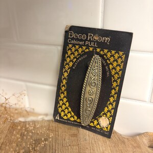 May include: A brass-toned metal cabinet pull with an elongated oval shape and embossed scrollwork, mounted on a black and yellow display card labeled Deco Room Cabinet PULL, RUSTPROOF, DURA-METAL, and American Tack & Hardware Inc., New York, N.Y.
