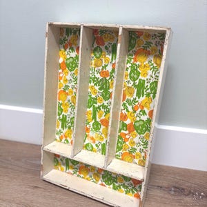 May include: A rectangular wooden cutlery tray with a distressed white painted finish. The interior base of the tray features a vibrant, repeating pattern of colorful vegetables, including orange onions, green artichokes, and leafy greens.