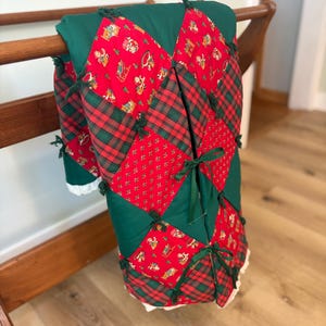 May include: A quilted blanket featuring a diamond-patterned patchwork design in red, dark green, and plaid fabric. The quilt is accented with small green fabric ties at the corners of the diamonds and finished with a white lace trim along the bottom edge.