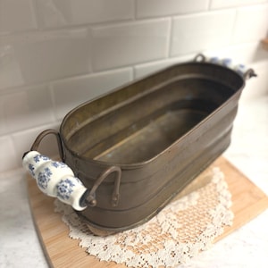 May include: An antique brass-toned metal planter with a rectangular shape and curved sides. The planter has two handles with white ceramic grips adorned with blue floral patterns. It rests on a wooden board with a lace doily.