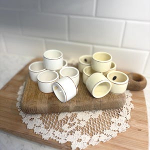 May include: A collection of white ceramic napkin rings with a gold trim, arranged on a wooden cutting board. Some rings are stacked, others are scattered. The background includes a white tile wall and a lace doily.