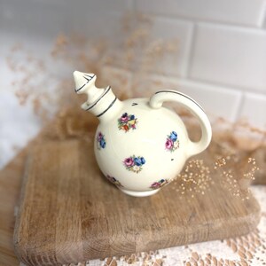 May include: A cream-colored ceramic oil cruet with a spherical body and a matching stopper. The surface is decorated with small, scattered floral clusters in shades of pink, blue, and green. It features a curved handle and a pointed top.