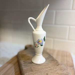 May include: A small, white ceramic vase with a handle and a flared spout. The vase features a painted floral design in shades of blue, pink, and green. It is displayed on a wooden cutting board. The vase is approximately 6 inches tall.
