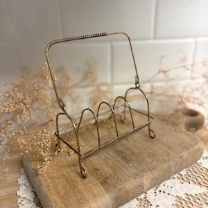 May include: A vintage-style brass wire toast rack featuring four arched slots for holding slices of bread and a single rectangular handle with a coiled wire grip. The metal structure has a weathered finish and stands on four small looped feet.