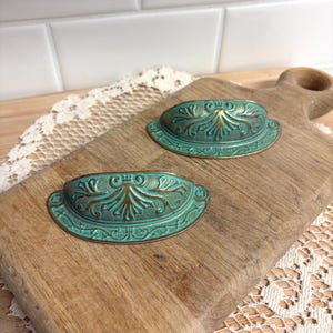 May include: Two antique-style drawer pulls with a teal and bronze finish. Each pull has an oval shape with ornate floral detailing. The pulls are displayed on a wooden cutting board, showcasing their vintage aesthetic.