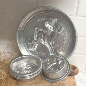 May include: A large round aluminum cake pan featuring a molded Rudolph the Red-Nosed Reindeer design with embossed text, accompanied by two smaller matching individual-sized aluminum molds, each also depicting the reindeer character.