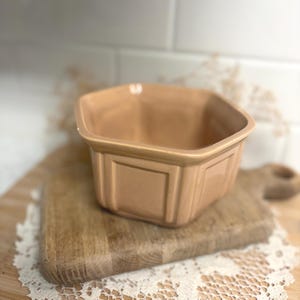 May include: A peach-colored ceramic bowl with a hexagonal shape and a smooth, glossy finish. The exterior features a raised, paneled design on each of the six sides, creating a structured appearance with a wide, open top and a flat base.
