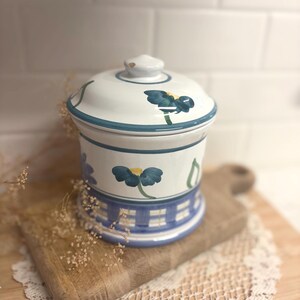 May include: A white ceramic canister with a matching lid featuring hand-painted dark blue flowers with yellow centers. The base of the canister is decorated with a blue and white checkered pattern, and the lid has a small knob handle on top.
