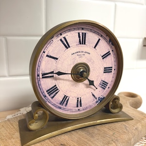 May include: Antique brass clock with a light pink face and black Roman numerals. The clock face reads "Baldwin and Jones, Benson, USA, 1872". The clock has a decorative base with scroll details.