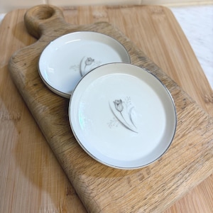 May include: Two small, round, white porcelain dishes with a silver rim and a gray tulip design. The dishes are placed on a wooden cutting board with a handle. The background is a light-colored wall.