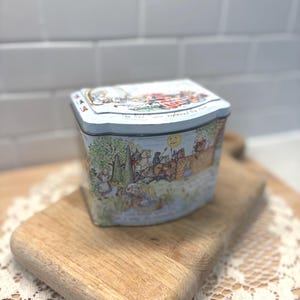 May include: A vintage, light blue tin box with a curved top and sides. The box features a colorful illustration of a story scene with text that reads "The Red Queen explained the rule." The tin is placed on a wooden cutting board.