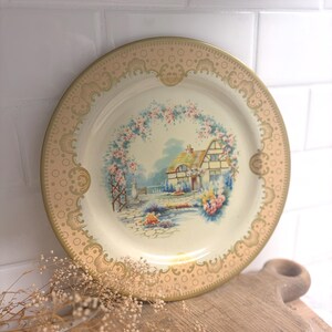 May include: A round decorative plate with a cream-colored center featuring a painted cottage scene surrounded by a floral arch. The rim is a wide, metallic gold band decorated with a repeating pattern of circles and scalloped, ornate motifs.