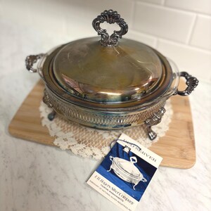 May include: Silver-plated serving dish with a lid and ornate handles. The dish sits on a wooden board with a lace doily. A tag from the E.B. Rogers Silver Company is visible, with the words "DUO SERVER".