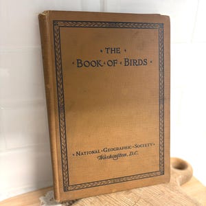 May include: A vintage book titled "The Book of Birds" with a tan cover and black text. The cover features a decorative border and the text "National Geographic Society, Washington, D.C."