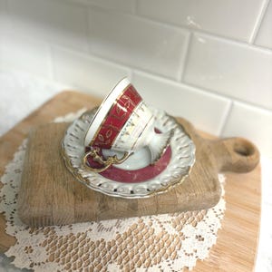 May include: An ornate teacup and saucer set. The teacup features a red band with gold accents and a gold handle. The saucer is white with a gold trim and a red center. The set is placed on a wooden cutting board.