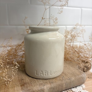 May include: A cream-colored ceramic jar with a smooth, glossy finish and a short, wide neck. The base of the jar features the embossed text "LAB Lagny" in capital and lowercase letters. The vessel has a cylindrical body with a slightly rounded rim.