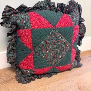 May include: A square quilted throw pillow featuring a central star pattern in red and dark green fabric with a holly berry print. The pillow is finished with a ruffled border made from the same holly-patterned fabric, creating a decorative edge.