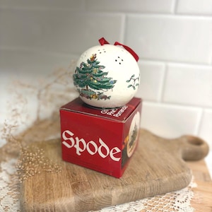 May include: A white ceramic spherical pomander with a Christmas tree illustration, featuring a red ribbon hanger and small ventilation holes. It sits atop a red cardboard box with the white text Spode printed on the front side.