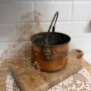 May include: A round copper pot with a dark patina, featuring a metal swing handle attached to the sides with decorative brass mounts. The pot has a smooth, cylindrical body with a horizontal band detail and a weathered interior surface.