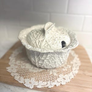 May include: A white ceramic cabbage-shaped serving dish with a lid, resting on a white lace doily and a wooden cutting board. The dish has a textured surface, and the lid features a handle. The overall theme is kitchenware.