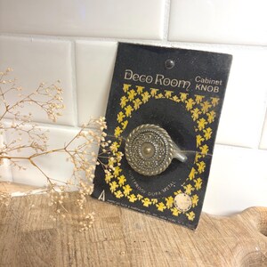 May include: A round, antique brass-toned metal cabinet knob with a rope-style border and intricate embossed center detail, mounted on a black and yellow Deco Room display card with the text Deco Room Cabinet Knob and Rustproof Dura Metal.