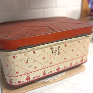 May include: Vintage rectangular tin with a red lid and cream-colored sides. The sides feature a red diamond pattern with small red flowers and Dutch figures. The tin sits on a wooden cutting board.