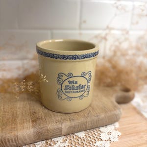 May include: A cylindrical beige stoneware crock featuring a blue decorative border around the rim and a blue graphic design on the front that reads Win Schuler RESTAURANTS enclosed within a stylized scrollwork frame with intricate detailing.