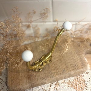 May include: A brass wall hook featuring two white ceramic ball finials at the ends of the curved metal arms. The base of the hook has an ornate, elongated shape with four visible screw holes for mounting to a flat surface.