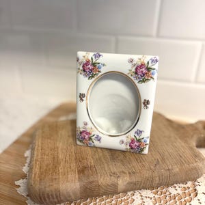 May include: A white porcelain picture frame with a gold trim around the oval opening. The frame is decorated with floral patterns in shades of pink, purple, and blue. The frame is displayed on a wooden cutting board.