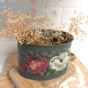May include: A cylindrical dark green metal container featuring hand-painted floral motifs in shades of deep red and white with green leaves. The container is filled with dried, light beige baby's breath flowers, creating a decorative arrangement.