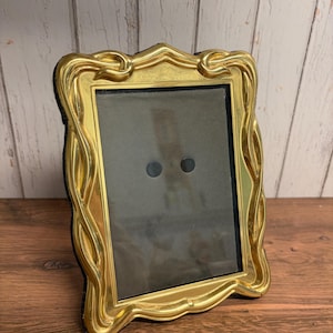 May include: A rectangular picture frame featuring a polished gold-tone metal border with an ornate, flowing, and wavy design. The frame contains a dark gray insert with two small, circular black magnets positioned in the center area.
