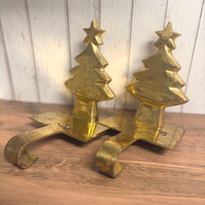 May include: A pair of polished gold-toned metal stocking holders, each featuring a stylized Christmas tree silhouette topped with a star. The base of each holder includes a flat rectangular plate and a curved hook for hanging.
