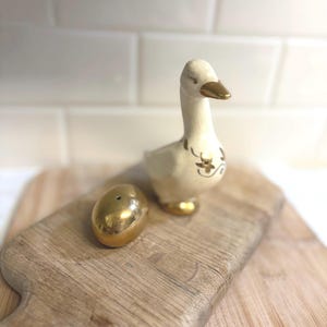 May include: A ceramic duck figurine with gold accents and a matching gold egg-shaped salt shaker. The duck is cream-colored with gold detailing on its wings and feet. The items are on a wooden cutting board.
