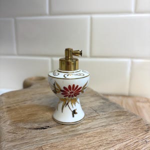 May include: A small, white ceramic perfume bottle with a gold-colored top and a red floral design. The bottle has a decorative shape and is placed on a wooden cutting board. The bottle is an antique or vintage item.