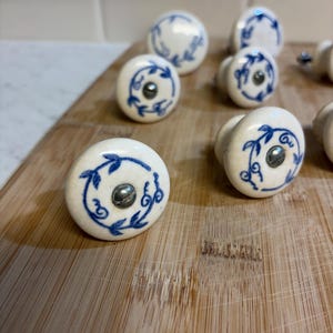 May include: A collection of white ceramic cabinet knobs with a blue floral design. Each knob features a silver screw and a rounded shape. The knobs are arranged on a wooden cutting board, showcasing their decorative style.