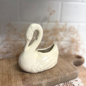 May include: A cream-colored ceramic swan planter featuring a glossy finish and molded feather details on the wings. The swan has a curved neck and an open cavity on its back for holding small items or plants, presented as a decorative object.