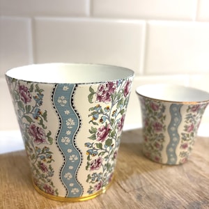 May include: Two ceramic planters with floral patterns. The larger planter has a cream background with a blue wavy stripe and pink and purple flowers. The smaller planter has a similar design. Both have gold rims.