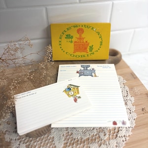May include: A yellow recipe box with the text Here's What's Cookin' and a vintage stove illustration. Included are matching lined recipe cards and a larger lined recipe sheet, both featuring printed kitchen-themed graphics and text.
