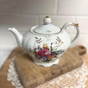 May include: White porcelain teapot with a floral design featuring pink, orange, and purple flowers. The teapot has a gold handle and trim, and a matching lid with a gold knob. It sits on a wooden cutting board.