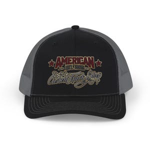 Puede incluir: Gorra de camionero negra y gris con el texto "AMERICAN EST. 2025 Custom Truck Shop" en burdeos y dorado. La gorra tiene una visera curva y una parte trasera de malla. Un accesorio elegante para los amantes de los camiones.