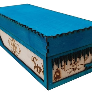Op de afbeelding: Een rechthoekige houten kist met een levendig blauwgroen deksel en zijkanten. De kist heeft ingewikkelde lasergesneden ontwerpen en tekst op de lichtgekleurde houten panelen. De kist heeft een decoratieve rand op het deksel en de zijkanten.