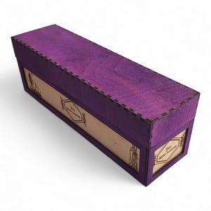 Puede incluir: Una caja rectangular de madera con exterior teñido de púrpura y paneles de madera natural. La caja presenta el texto "THE RING-MAKER" en un marco decorativo. La caja tiene un diseño ranurado en los bordes.